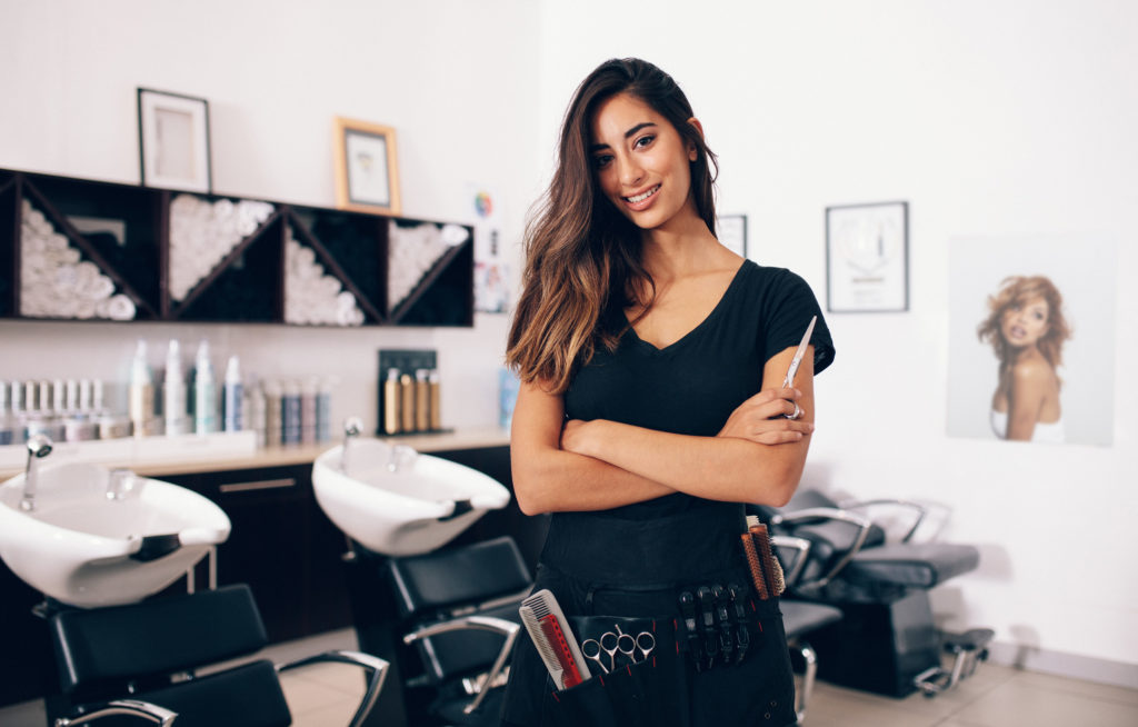 Vrouw staat met armen over elkaar in kapsalon en kijkt in camera